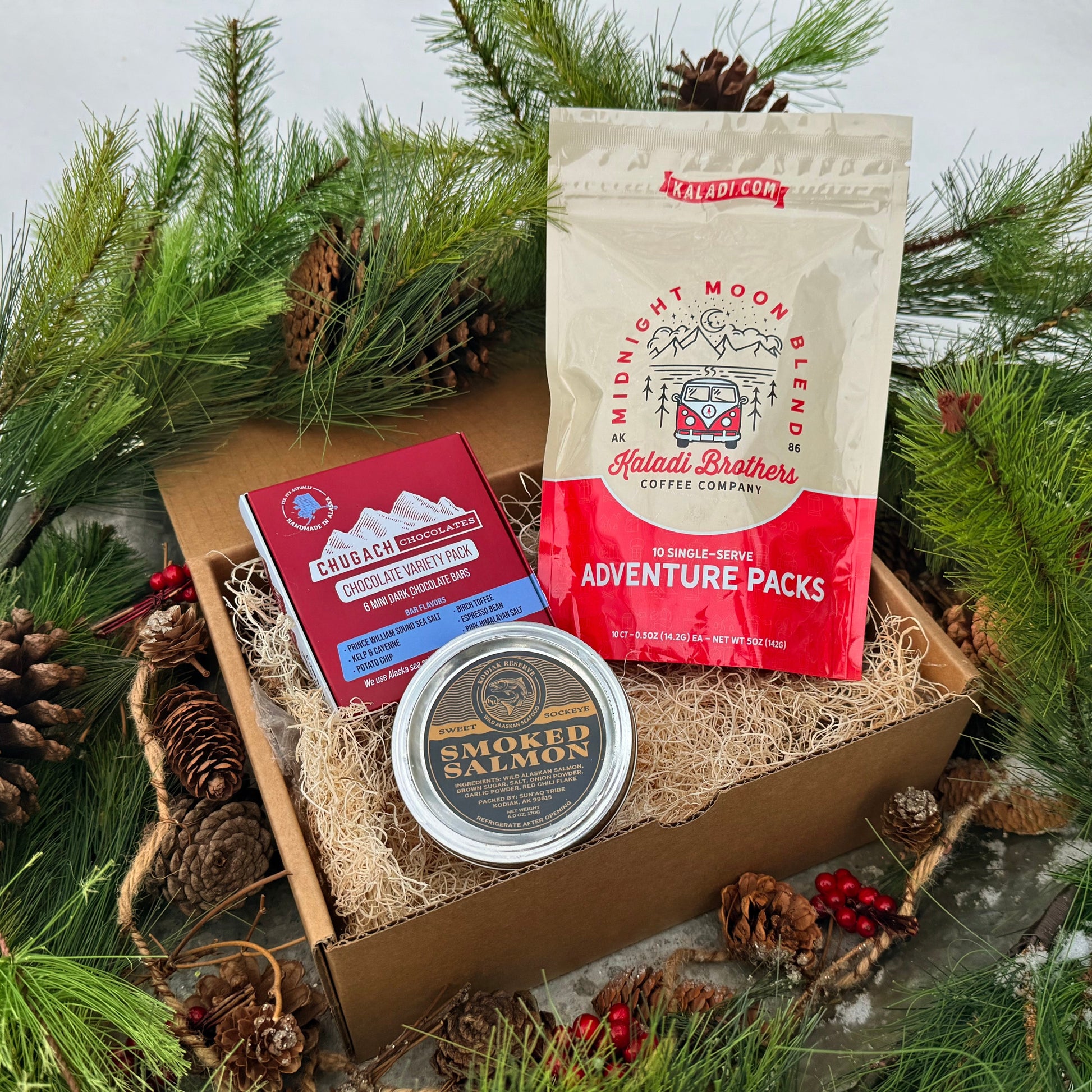 Gift box with salmon jar, coffee, and chocolate variety pack surrounded by pine branches and cones.