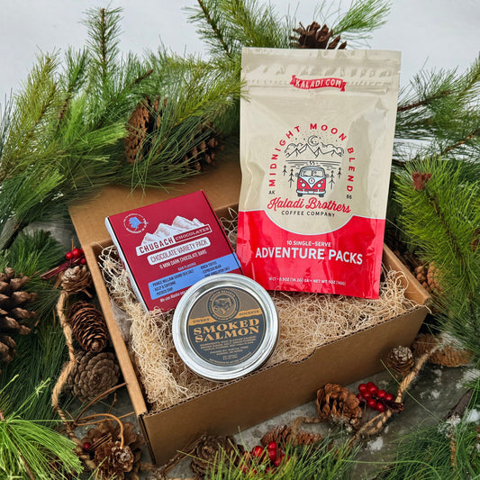 Gift box with salmon jar, coffee, and chocolate variety pack surrounded by pine branches and cones.