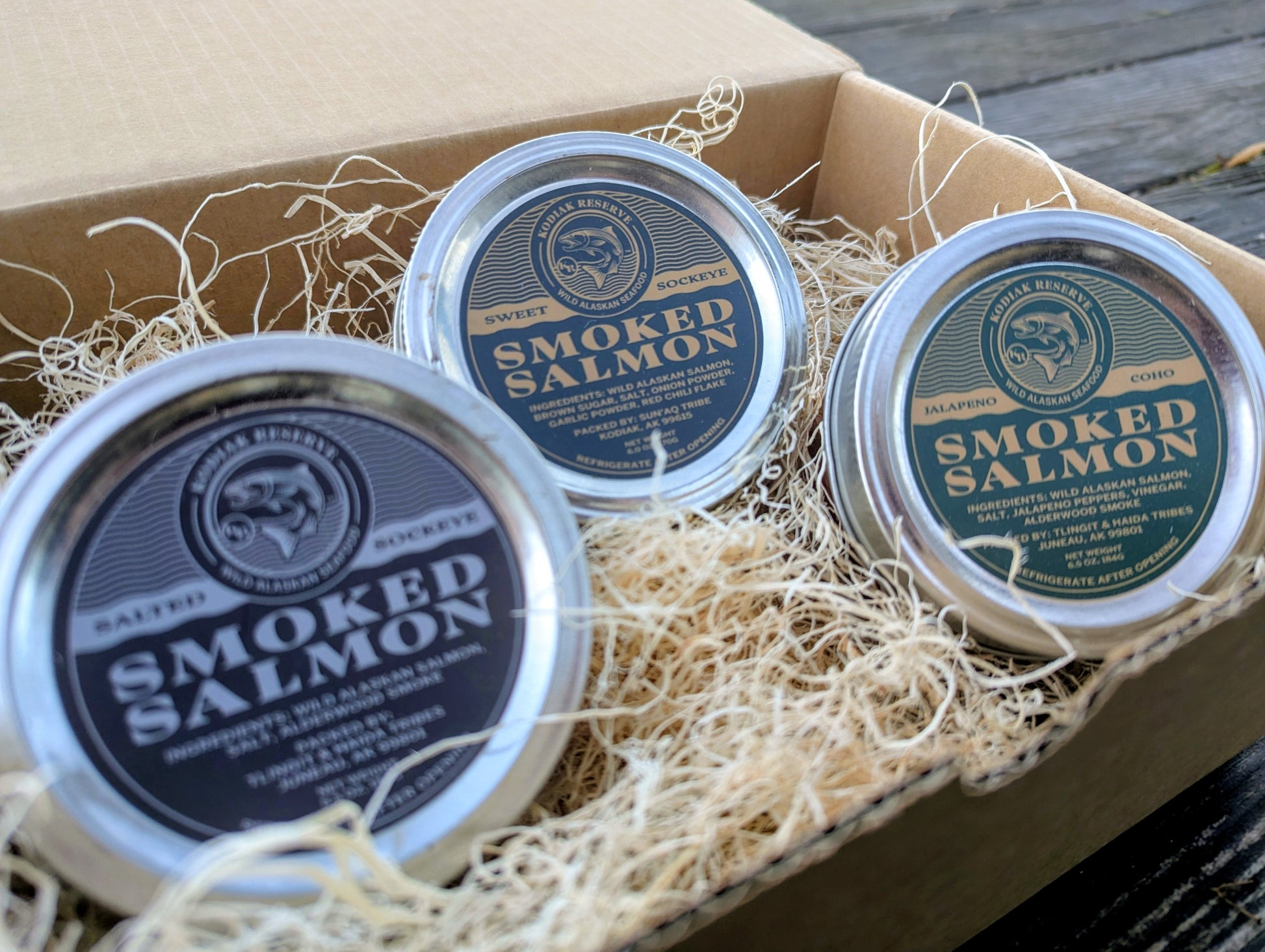 Three tins of smoked salmon in a cardboard box with decorative Aspen