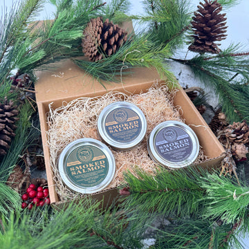 Three tins of Smoked Salmon in a decorative box with pine branches and cones.